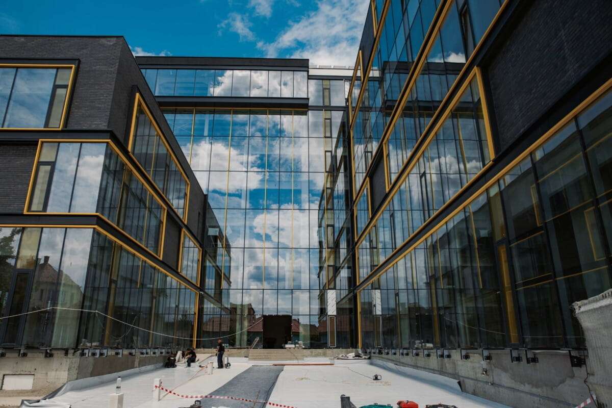 The building reflects the sky vividly, highlighting the seamless integration of performance-focused glazing. In the foreground, ongoing waterproofing and insulation work illustrates the meticulous attention to fire safety, water resistance, and acoustic control integral to modern façade design